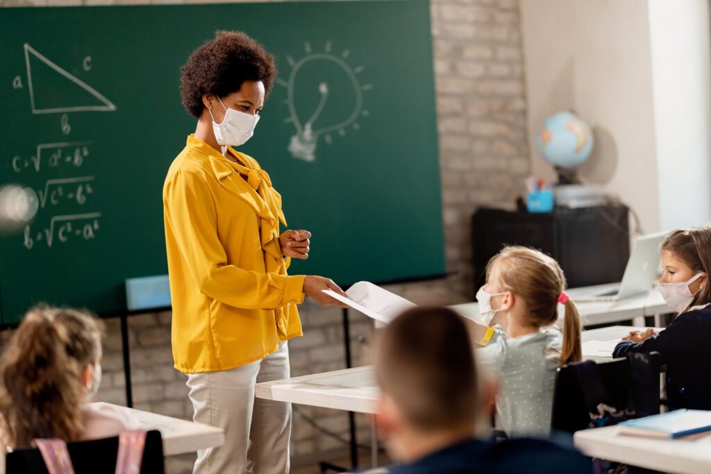 Happy black teacher and her students wearing protective face mask in the classroom. Teacher is giving them their test results. (Happy black teacher and her students wearing protective face mask in the classroom. Teacher is giving them their test resu